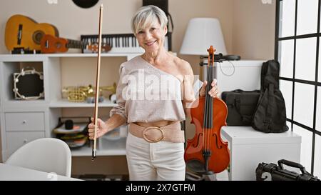 Donna matura con capelli grigi che tiene il violino e l'arco in uno studio musicale circondato da vari strumenti. Foto Stock