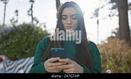 Giovane donna ispanica che utilizza lo smartphone su strade urbane soleggiate con retroilluminazione naturale e alberi sullo sfondo. Foto Stock