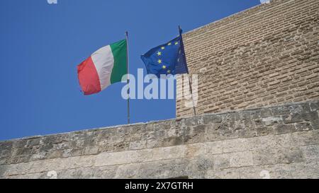 Bandiere italiane ed europee che sventolano contro un cielo azzurro e limpido in cima a uno storico edificio in pietra che rappresenta l'italia, l'europa e l'unità internazionale. Foto Stock