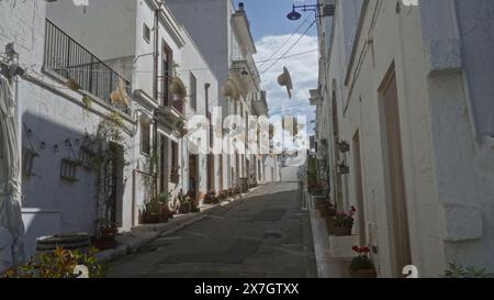 Affascinante strada ad alberobello, italia, puglia, decorata con cappelli appesi, fiori e piante in vaso sotto un cielo blu in un ambiente storico della città. Foto Stock