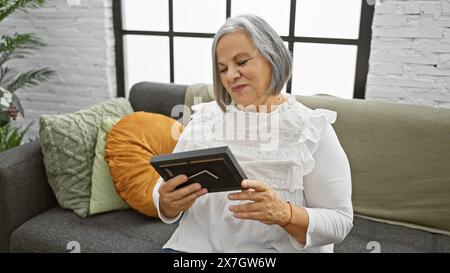 Una donna sorridente di mezza età legge un tablet in un salotto accogliente, incarnando comfort e tecnologia moderna a casa. Foto Stock