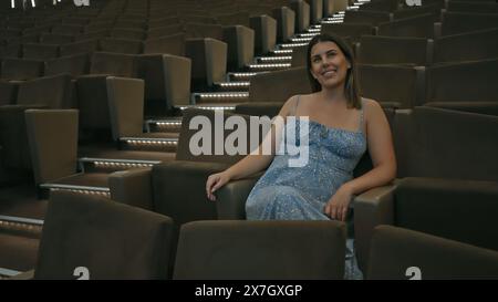 Una giovane donna ispanica seduta da sola in un teatro buio e vuoto, che sembra allegra e rilassata. Foto Stock