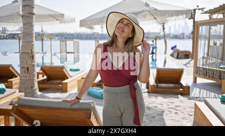 Una donna sorridente con cappello da sole e abbigliamento da spiaggia gode dell'ambiente lussuoso di un'area piscina di lusso sulla spiaggia. Foto Stock