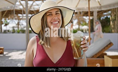 Una donna sorridente in un cappello da sole gode di un mojito in un resort sulla spiaggia di lusso, che rappresenta il relax e il tempo libero. Foto Stock