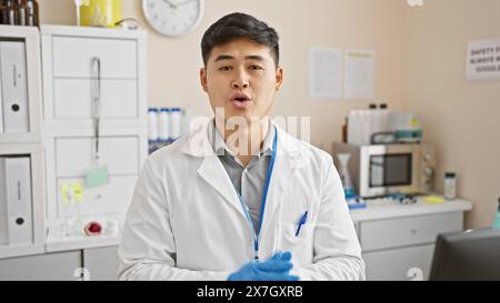 Scienziato maschio asiatico in camice da laboratorio in piedi in laboratorio con attrezzature e poster di sicurezza in background Foto Stock