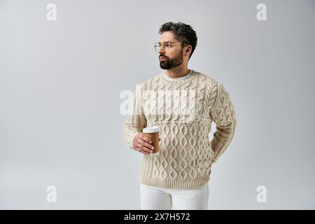 Un uomo con barba trasuda fascino con un pullover bianco su sfondo grigio, che sfoggia un look sofisticato e raffinato. Foto Stock