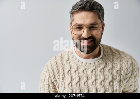 Un uomo con barba trasuda fascino con un maglione bianco, completato da occhiali, su uno sfondo grigio in un ambiente da studio. Foto Stock