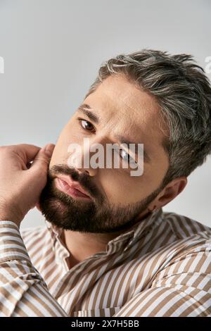 Un uomo elegante con una barba che trasuda fascino in una camicia a righe su uno sfondo da studio neutro. Foto Stock