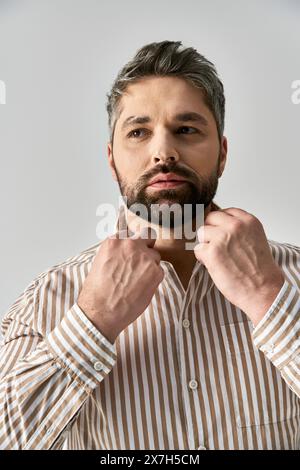 Un uomo con barba trasuda fiducia in un'elegante camicia a righe su sfondo grigio studio. Foto Stock