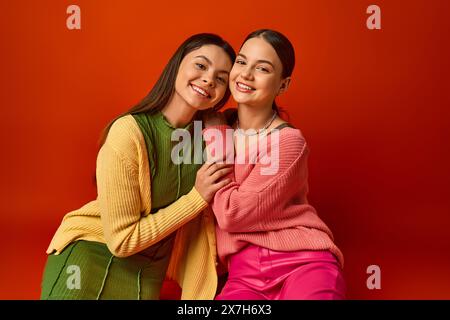 Due graziose ragazze brune in un abbigliamento casual sedute l'una accanto all'altra su un vivace sfondo rosso in un ambiente studio. Foto Stock