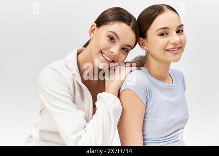 Due simpatici amici brune in abbigliamento sportivo si siedono fianco a fianco, condividendo sorrisi e felicità su uno sfondo grigio. Foto Stock