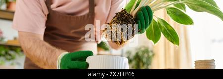 Un uomo con una camicia rosa e guanti verdi tiene delicatamente una pianta in un accogliente negozio di piante, incarnando l'essenza della cura dei fioristi. Foto Stock