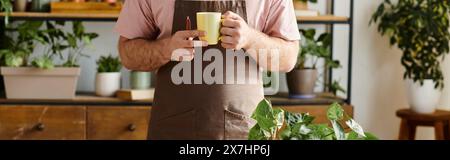 uomo corto che tiene in mano una tazza mentre si occupa delle piante nel suo piccolo negozio di piante. Foto Stock