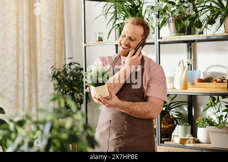 Un uomo elegante multitasking, conversando su un cellulare mentre tiene delicatamente una pianta in vaso in un negozio di piante. Foto Stock