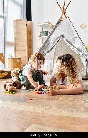 Una madre dai capelli ricci e la figlia del bambino giocano felicemente con i giocattoli sul pavimento, praticando il metodo educativo Montessori. Foto Stock