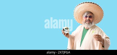 Ritratto di un uomo maturo felice in poncho e sombrero che tiene in mano tequila e teschio dipinto su sfondo blu con spazio per il testo Foto Stock