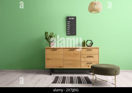 Cassettiera in legno con arredamento e pouf vicino alla parete verde in un elegante interno del soggiorno Foto Stock