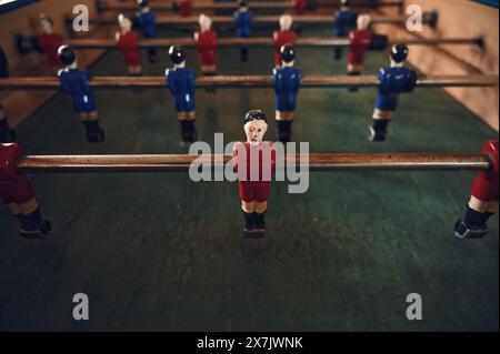 Primo piano di un giocatore di foosball dalla faccia pallida con un'uniforme rossa, fortemente a fuoco contro una fila di avversari blu sfocati, su un campo verde profondo e ben usurato. Foto Stock