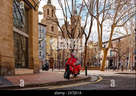 Scooter classico rosso parcheggiato in un angolo da una piazza alberata senza fronzoli con la chiesa di Santa Isabel de Portugal in stile barocco. Foto Stock