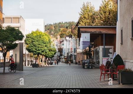 La gente passeggia lungo la strada pedonale principale di Vaduz, Liechtenstein, fiancheggiata da negozi ed edifici, con il fogliame autunnale sullo sfondo Foto Stock