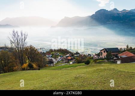 Un villaggio collinare nel Liechtenstein con vista sulla valle sottostante, avvolto dalla nebbia in un pomeriggio limpido Foto Stock