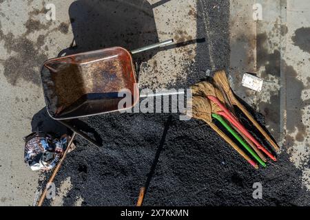 Una carriola vuota e attrezzi vicino a un mucchio di asfalto schiacciato adagiato sulla strada Foto Stock