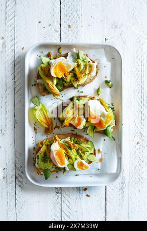 Fiori di zucchine fritti su fette di baguette, serviti con uova in camicia e erba cipollina su fondo bianco di legno Foto Stock