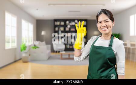 La governante asiatica indossa guanti di gomma gialli e grembiule verde che sorride nel soggiorno Foto Stock