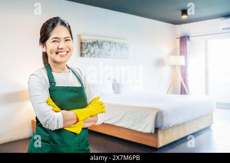 La governante asiatica indossa guanti di gomma gialli e grembiule verde che sorride in camera da letto Foto Stock