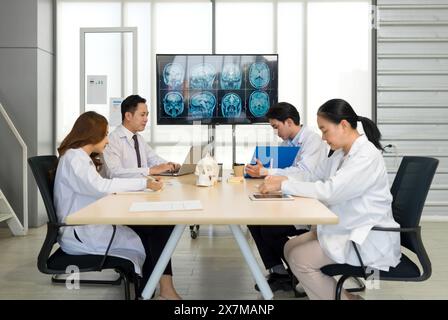 Gruppo di medici professionisti impegnati in una discussione o riunione in uno studio medico. Un monitor di grandi dimensioni che visualizza più scansioni cerebrali RM, per ruotare Foto Stock