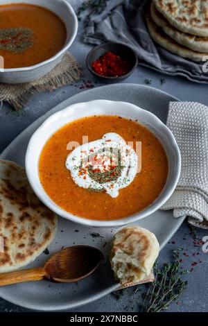 Zuppa di Tarhana ricoperta di yogurt, menta secca e peperoni rossi in un recipiente bianco, pane piatto e un cucchiaio di legno accompagnano. Foto Stock