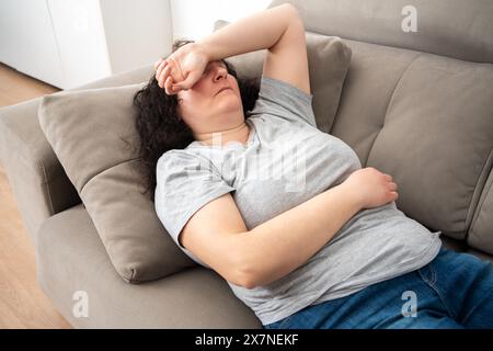 Donna sdraiata sulla schiena sul comodo divano di casa, che copre gli occhi con la mano, dorme di giorno, si sente stanca, si gode il relax, il tempo libero, pausa, noi Foto Stock