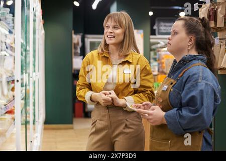 Ritratto di un manager sorridente che sta addestrando una giovane donna con sindrome di Down che lavora al supermercato e guarda l'esposizione del congelatore Foto Stock