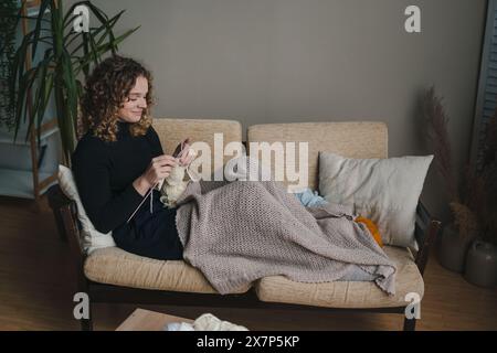 Giovane donna caucasica che lavora a maglia a mano mentre si siede sul divano a casa e si gode un accogliente hobby. Foto Stock