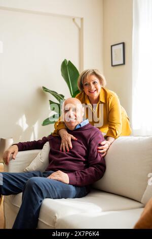 Una coppia anziana sorridente insieme in un soggiorno accogliente e soleggiato. La donna sta dietro l'uomo, abbracciandolo affettuosamente mentre siede su una sof Foto Stock