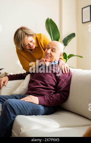 Una coppia anziana sorridente insieme in un soggiorno accogliente e soleggiato. La donna sta dietro l'uomo, abbracciandolo affettuosamente mentre siede su una sof Foto Stock