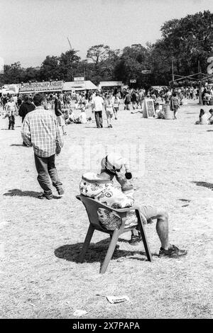 CROWD, HEATWAVE, NME STAGE, GLASTONBURY 95: Un uomo riposa su una sedia di plastica in un cappello bobble di legno sull'erba durante l'estrema ondata di caldo estiva presso le bancarelle di cibo al Glastonbury Festival, Pilton Farm, Somerset, Inghilterra, 24 giugno 1995. Nel 1995 il festival ha celebrato il suo 25 ° anniversario. Molte persone hanno lottato con il colpo di calore nel weekend particolarmente caldo. Foto: ROB WATKINS Foto Stock