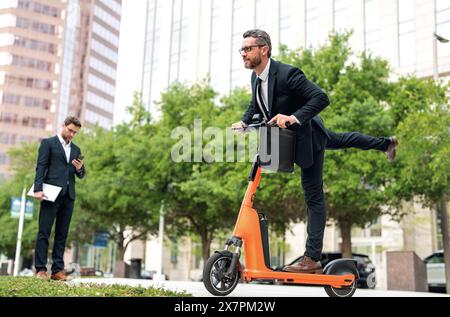 Un uomo d'affari eccitato e divertente su uno scooter elettrico per le strade cittadine. Impiegato uomo d'affari in tuta avvocato aziendale in tuta che guida e-scooter. Uomo d'affari Foto Stock