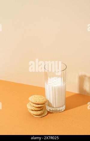 Una pila ordinata di biscotti croccanti con farina d'avena accanto a un bicchiere pieno di latte su una superficie arancione, con ombre morbide. Foto Stock