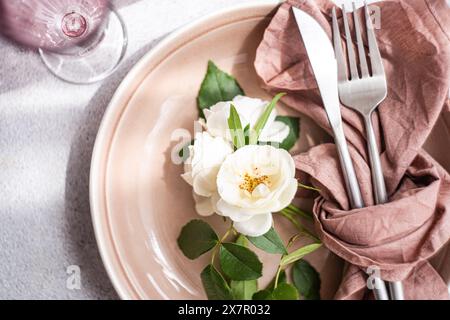 Un sofisticato tavolo con un piatto di ceramica pallida, posate in acciaio inossidabile avvolte in un tovagliolo di lino rosa scuro e una disposizione delicata Foto Stock