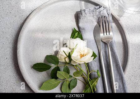 Un sofisticato tavolo con piatto in ceramica neutra, posate in argento, tovagliolo in lino e una disposizione di rose bianche fresche, ideale per matrimoni Foto Stock