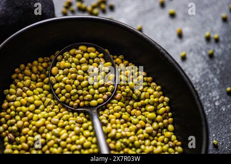 Una foto ravvicinata che mostra i fagioli mung biologici grezzi in una padella scura, con un cucchiaio di fagioli in mezzo a una disposizione sparsa Foto Stock