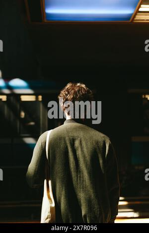 Un giovane uomo sta con le spalle alla telecamera, inghiottito di ombra, mentre una luce brillante illumina la parte superiore di una scena urbana dietro di lui il co Foto Stock