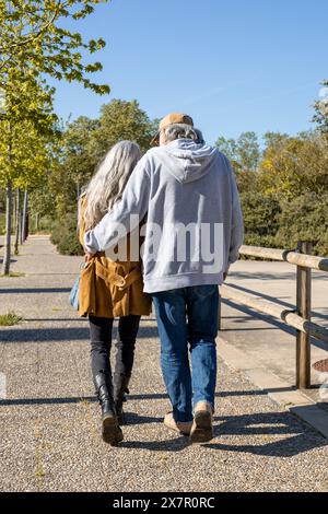Un'affettuosa coppia di anziani cammina di mano lungo un sentiero soleggiato del parco, mostrando un bellissimo momento di compagnia e amore nei loro anni d'oro Foto Stock