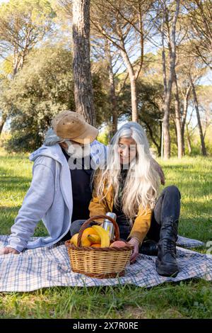 Una coppia senior siede comodamente su una coperta da picnic in un parco lussureggiante, circondato da alberi, condividono un momento di gioia insieme, esaminando la frutta Foto Stock