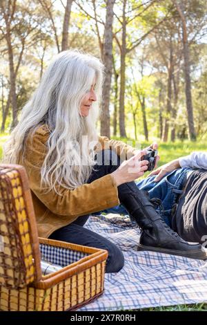 Una donna anziana con lunghi capelli grigi spara affettuosamente al suo partner mentre è seduta su una coperta da picnic in un parco lussureggiante, una scena idilliaca che cattura un uomo Foto Stock