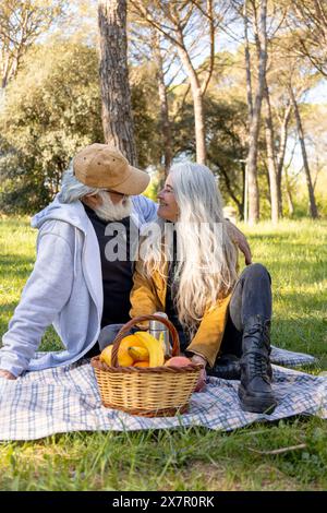 Una coppia senior condivide un momento di amore durante un picnic in un parco lussureggiante, circondato da alberi, con un cestino di frutta accanto Foto Stock