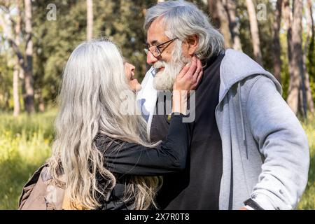 Un'affettuosa coppia senior che si guarda in un parco illuminato dal sole, con l'uomo che culla dolcemente il volto della donna, circondato da una vegetazione lussureggiante Foto Stock