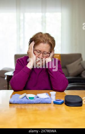 Una donna anziana si siede a un tavolo sentendosi afflitta e tenendo la testa, con strumenti di gestione del diabete davanti a lei, che illustrano i sintomi dell'ipog Foto Stock