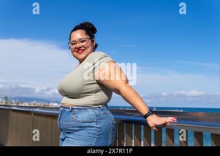 Una donna allegra e di grandi dimensioni sorride mentre ama una giornata estiva con gli amici su un balcone di Barcellona, con un cielo azzurro e lo sfondo dell'oceano Foto Stock